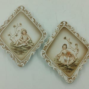 May include: Two white ceramic wall plaques with ornate frames. Each plaque features a raised relief image of a child in a woodland setting. One plaque depicts a boy sitting on a tree stump, while the other plaque depicts a girl standing in a forest.