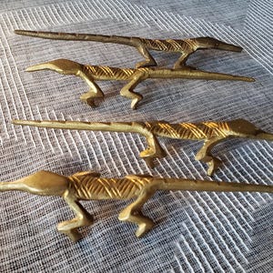 May include: Four brass lizard figurines arranged in a vertical row. Each lizard features a metallic gold finish with etched diagonal line patterns along the body and tail, four legs, and a pointed snout, showcasing a consistent design across all pieces.