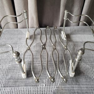 May include: A silver-toned metal expandable rack featuring a series of interconnected, scissor-like folding arms. The structure includes four vertical supports with curved wire frames and small white plastic components at the base of each support.