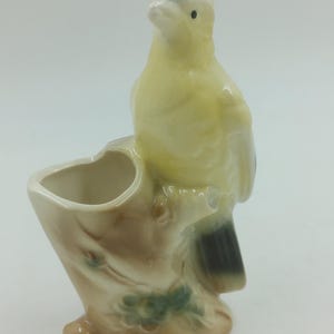 以下が含まれることがあります： 黄色、黒、茶色のセラミック製の鳥の置物。鳥は、植木鉢または花瓶の可能性がある小さな開口部のある木の幹にとまっています。置物は光沢のある仕上がりです。