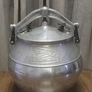 May include: A silver pressure cooker with a large handle and a screw-down mechanism. The pot has a rounded base and a lid with a locking system. The front of the pot has an embossed design with text.