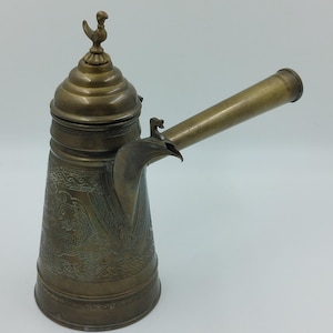 May include: A brass coffee pot with an ornate design and a bird finial on the lid. The pot has a long spout and a handle.
