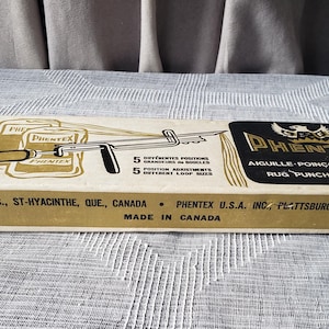 May include: A rectangular cardboard box for a Phentex rug punching needle. The box features a graphic of the tool, text stating 5 different loop sizes, and the brand name Phentex. It includes company information for St-Hyacinthe, Canada and Plattsburgh, N.Y.