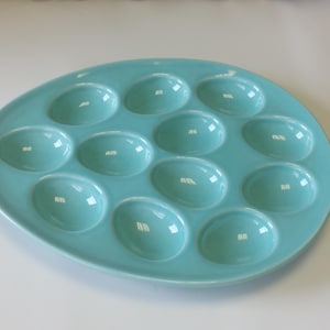 Peut inclure: Un plat de service à œufs en céramique turquoise avec 12 empreintes ovales. Le plat a une finition brillante et est conçu pour contenir des œufs pour le service. Le plateau à œufs est de couleur bleu clair et de forme ovale.