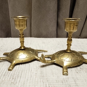 May include: Two brass candle holders in the shape of turtles. Each has a cup for a candle and a turtle-shaped base with legs and a shell. The candle holders are gold-coloured and sit on a light-coloured surface.