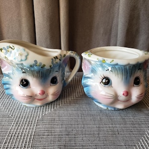 May include: A ceramic creamer and sugar bowl set featuring a blue and white cat face design. Each piece is painted with blue fur details, pink cheeks, and small blue flowers along the top rim. The creamer has a handle and a pouring spout.