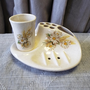 May include: A vintage ceramic bathroom set with a speckled cream finish. The set includes a toothbrush holder, soap dish, and a tumbler. Floral daisy and wheat sprig designs decorate the set. A charming addition to any bathroom.