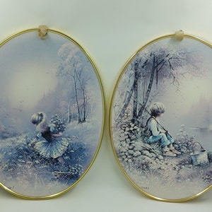 May include: Two framed oval paintings with a wintery theme. The left painting shows a girl in a blue dress picking flowers in a snowy forest. The right painting shows a boy in blue overalls sitting by a lake with a fishing rod.