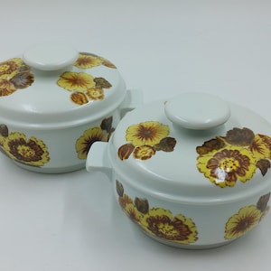 May include: Two white ceramic casserole dishes with floral designs. The dishes feature brown and yellow flowers with green leaves. The lids are white with a knob on top.