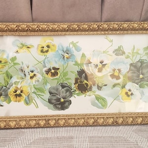 May include: A long, rectangular framed print featuring a horizontal arrangement of pansy flowers in shades of yellow, blue, white, and dark purple with green leaves. The frame is ornate, metallic gold, and has a textured, repeating pattern along its edge.