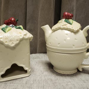 May include: A cream ceramic tea set and a matching rectangular container. The tea set includes a teapot with a lid and a teacup. Both the teapot lid and the container top are decorated with a strawberry and leaf design.