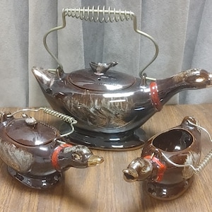 May include: A vintage brown ceramic duck-shaped tea set. The set includes a teapot with a metal handle, a sugar bowl, and a creamer. Each piece has a red collar detail and gold-coloured beak.
