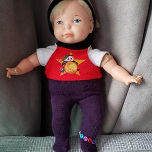 以下が含まれることがあります： ブロンドの髪、青い目、赤いリボンをしたヴィンテージドール。パンダのグラフィックが入った赤いシャツ、紫色のズボン、ビーズのアンクレットを着用しています。人形の両腕は広げられ、灰色の背景にポーズをとっています。