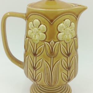 May include: A vintage ceramic jug in a warm, mustard yellow hue. The jug features a lid and handle, with a floral and leaf pattern embossed on the body. The base is a pedestal design.