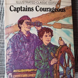 May include: A paperback book titled Captains Courageous by Rudyard Kipling, part of the Illustrated Classic Editions series. The cover features a color illustration of a person using a telescope and another person steering a ship's wooden wheel.