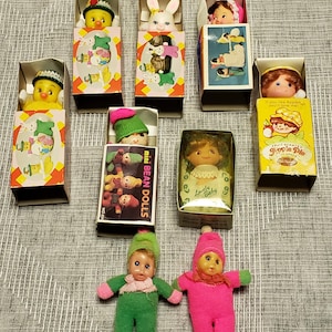 May include: A selection of vintage miniature dolls, including a chick, bunny, and baby dolls, presented in their original boxes. The boxes feature colourful illustrations and text, with two small dolls standing in front.