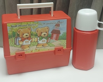 May include: A vintage red lunchbox and matching flask. The lunchbox features a graphic of two teddy bears and the text "Kraft Peanut Butter... the Peanuttiest Taste!". The flask has a white lid and handle.