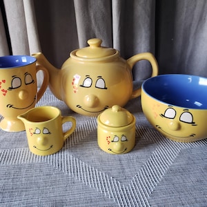 May include: A yellow ceramic tea set comprising a teapot, a mug with a blue interior, a creamer, a sugar bowl with a lid, and a bowl. Each item features a smiling face with closed eyes, a round nose, and small red heart accents on the cheeks.