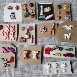 Peut inclure: Une collection de divers embellissements décoratifs, notamment des formes d'animaux, des chaussures, des rubans et des véhicules. Certains embellissements sont sur des cartes avec le mot "COOKIES" visible. Les couleurs incluent le rose, le rouge, le marron et le blanc.