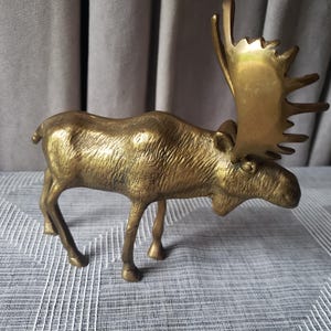 May include: A polished brass figurine of a standing moose. The sculpture features a textured body, a downward-facing head, and large, prominent antlers with multiple points. The metal surface has a warm, reflective golden tone throughout.