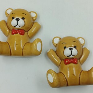 May include: Two ceramic teddy bear figurines with red bow ties. The bears are sitting with their arms raised and have white accents on their paws.