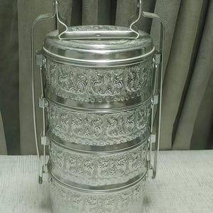 May include: A silver-coloured, tiered food carrier with an ornate floral pattern. The carrier has a handle on top and side handles for easy transport. The stacked containers are held together by a metal frame.
