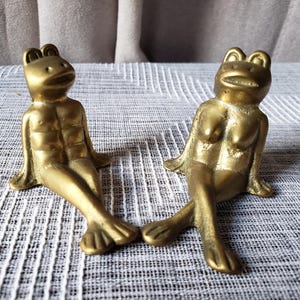 May include: Two brass frog figurines in a seated position. The left frog features a defined chest and torso, while the right frog features a defined chest. Both figures have smooth, metallic gold-toned surfaces, wide eyes, and webbed feet.
