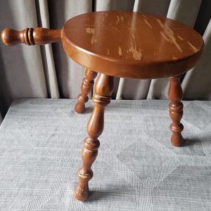 May include: A brown wooden stool with a round top and three turned legs. The stool features a long, horizontal handle extending from the side of the seat. The wood finish shows visible signs of wear, including scratches and scuffs on the surface.