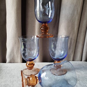 May include: Four blue and amber-coloured stemmed glasses. The blue glass has a floral design etched into the surface. The base and stem are amber-coloured. One glass is lying on its side. The glasses are on a white surface.