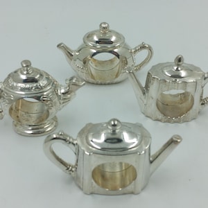 May include: Four silver teapot napkin rings. The rings are detailed with a teapot design and have a hole in the centre for a napkin.