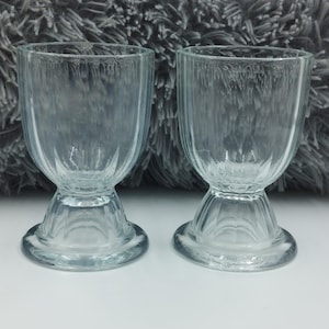 May include: Two clear glass shot glasses with a fluted design and a wide base. The glasses are sitting on a grey fluffy surface.