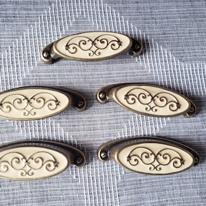 May include: Five oval-shaped drawer pulls featuring a cream-coloured centre inlay with an embossed bronze-toned metal scrollwork design. Each handle has a metallic bronze finish on the outer frame and includes two visible screw attachment points.