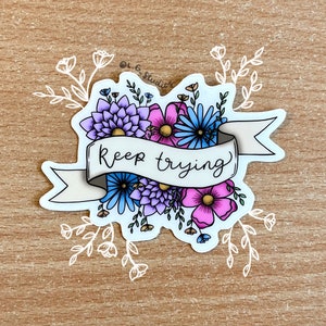 Op de afbeelding: Een sticker met een bloemenontwerp en de tekst "Keep trying". De bloemen zijn in tinten paars, blauw en roze.