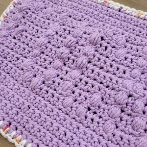 Peut inclure: Un gros plan sur un tapis crocheté violet avec une surface texturée et bosselée. Le tapis a une bordure blanche et orange.