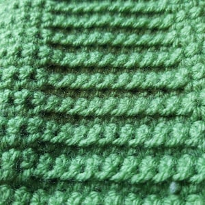 May include: Close-up of a green knitted fabric with a textured pattern. The fabric is made of yarn and has a soft, fuzzy texture.