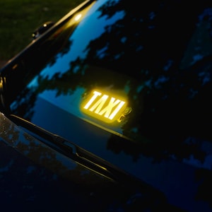 May include: A bright yellow illuminated taxi sign attached to the interior of a vehicle windshield. The sign features the word TAXI in bold, capitalized, sans-serif lettering, glowing against the dark glass of the car window at night.