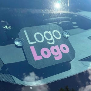 May include: A black square sign with rounded corners attached to a car windshield with two clear suction cups. The sign features the word Logo in white text on the top line and the word Logo in pink text on the bottom line, both in a sans-serif font.