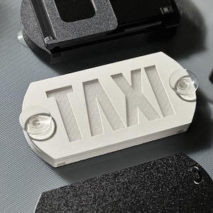 May include: White plastic sign with the word "TAXI" cut out in the center. The sign has two clear suction cups on the back.