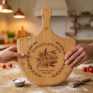 Może przedstawiać: Jasnobrązowa łopata do pizzy z bambusa z laserowo grawerowanym wzorem kawałka pizzy otoczonego wieńcem laurowym. Napis głosi SOPHIA'S KITCHEN oraz MADE WITH LOVE SINCE 2021. Deska posiada długi uchwyt z otworem w górnej części.