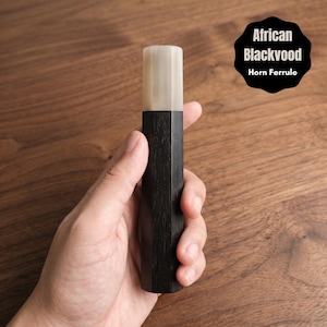 May include: A dark, hexagonal wooden object with a light-colored top, held in a hand. The object is labeled "African Blackwood Horn Ferrule" in a black circular graphic.