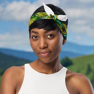 This is an image of a woman wearing the green mushroom scarf tied around her head as a headband standing in front of a mountain range.