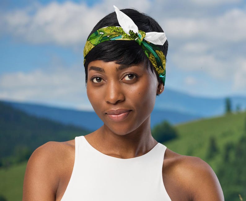 This is an image of a woman wearing the green mushroom scarf tied around her head as a headband standing in front of a mountain range.