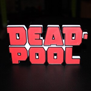Op de afbeelding: Rood en wit "DEADPOOL" logo op een zwarte ondergrond. De letters zijn rood met een witte rand. De achtergrond is donker. Het logo is van een strip- en filmkarakter.
