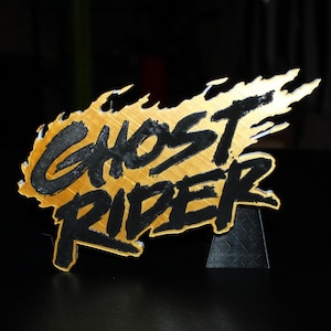 Pode incluir: Uma placa dourada com as palavras "GHOST RIDER" em preto, com um design em forma de chama. A placa está montada em um suporte preto. O tema geral está relacionado a uma banda desenhada ou filme.