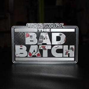 May include: A rectangular Star Wars "The Bad Batch" sign with a silver border and lettering. The text is in a distressed style with red accents. The sign is on a dark surface.