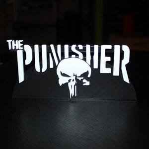 May include: A black and white sign featuring the text "THE PUNISHER" above a skull logo. The sign is made of a dark material and is likely a decorative item or collectible.