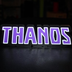 May include: A glowing sign with the name "THANOS" in purple letters outlined in white. The sign has a black border and is illuminated against a dark background. The letters are bold and easy to read.