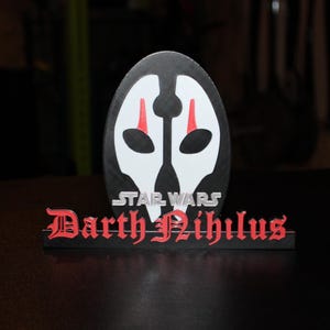 Op de afbeelding: Een zwart-wit Darth Nihilus Star Wars-karaktermasker met rode accenten. Het Star Wars-logo staat boven het masker en de naam "Darth Nihilus" staat in rode gotische letters eronder.