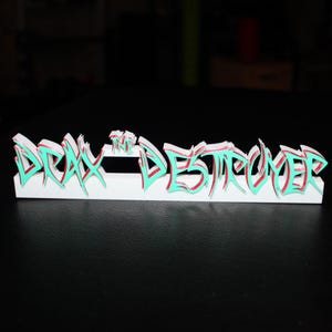 Puede incluir: Un letrero blanco impreso en 3D con el texto "DRAX THE DESTROYER" en una fuente de estilo grafiti. El texto está delineado en verde azulado y rojo, con un efecto de sombra. El letrero está sobre una superficie negra.