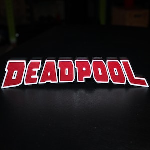 May include: A red and white "DEADPOOL" logo, featuring a bold, blocky font. The letters are red with a white outline, set against a dark surface. This logo is likely a collectible or decorative item.